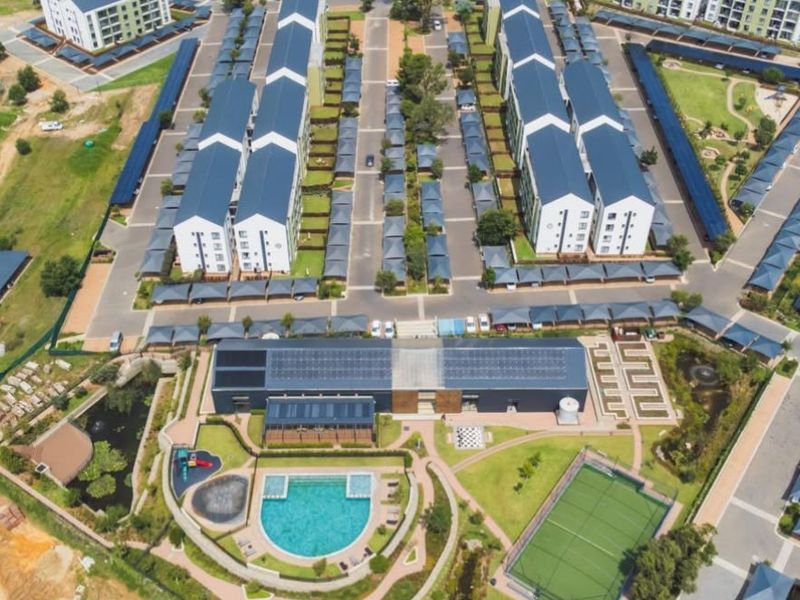 Aerial view of a modern apartment complex featuring multiple residential blocks, covered parking, a swimming pool, tennis court, and communal clubhouse facilities.