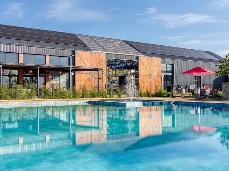 A modern communal pool area featuring a large swimming pool with a water fountain, set against a contemporary building with solar panels and industrial-style architecture.