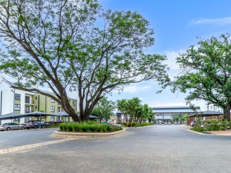 A well-maintained apartment complex entrance featuring a paved driveway, mature trees, landscaped garden beds, and covered parking bays.