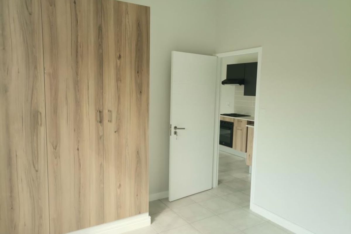 A bright, empty bedroom featuring modern wood-finish built-in wardrobes, neutral tiled flooring, and a view into the adjacent kitchen area.