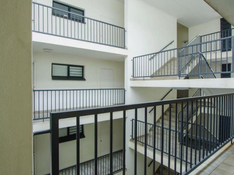 An open-air apartment complex hallway featuring metal balustrades, tiled walkways, and a central staircase providing access to multiple levels.