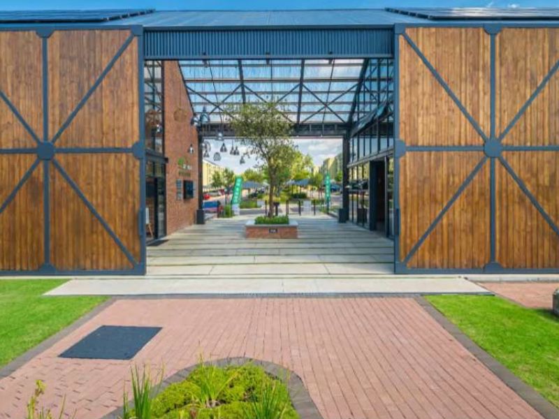 Modern apartment complex entrance featuring large timber sliding doors, industrial metal roofing, and a well-maintained paved approach with landscaped garden beds.