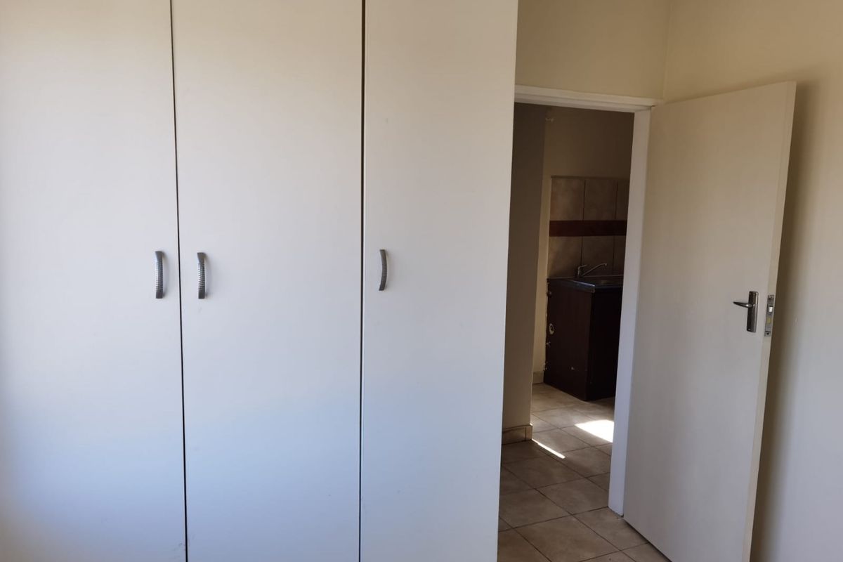 A clean, empty bedroom featuring built-in cupboards and tiled flooring, with a view through the doorway into an adjacent tiled utility or kitchen area.