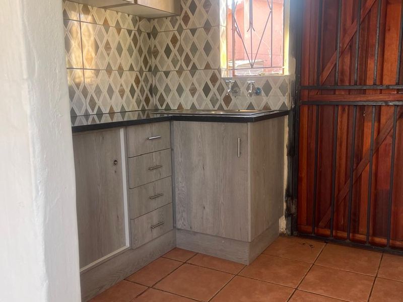 A compact kitchen featuring wood-look cabinetry, a stainless steel sink, patterned tiled backsplash, and terracotta floor tiles with an integrated security door.