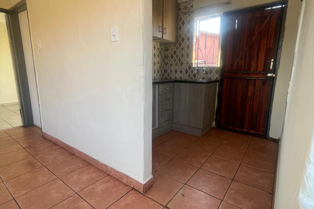 A compact kitchen area featuring built-in cabinetry, tiled backsplash, and terracotta-style floor tiles, with direct access to an exterior security door.