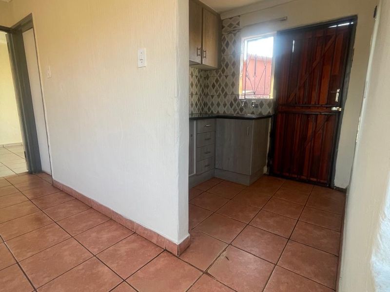 A compact kitchen area featuring built-in cabinetry, tiled backsplash, and terracotta-style floor tiles, with direct access to an exterior security door.