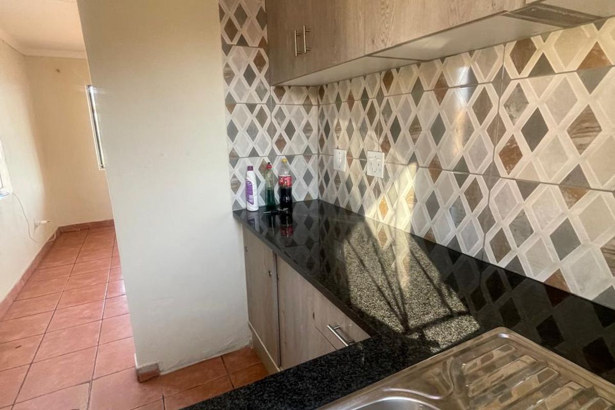 A functional kitchen area featuring granite countertops, wood-look cabinetry, and a patterned tile backsplash, leading into an adjacent tiled living space.