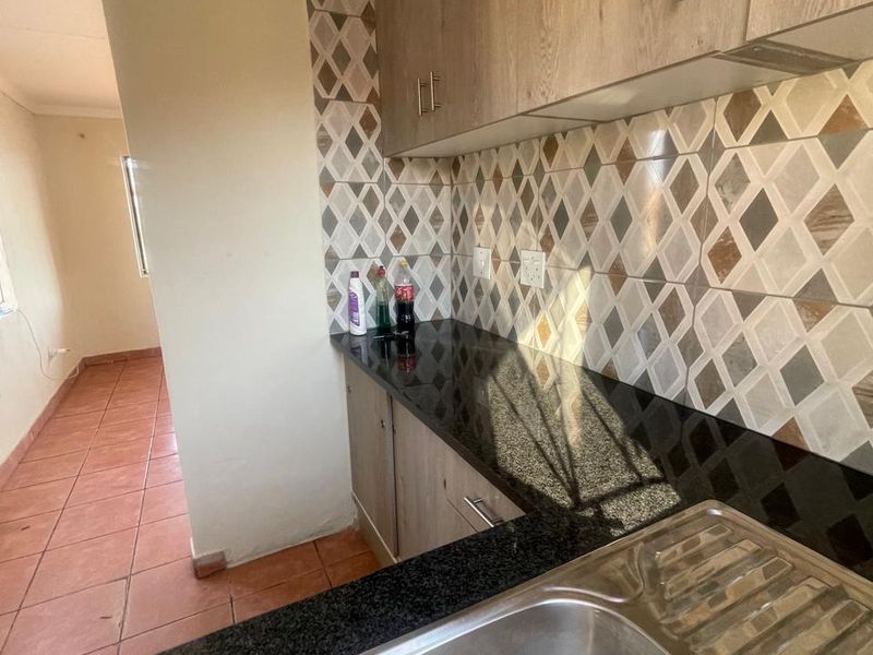 A functional kitchen area featuring granite countertops, wood-look cabinetry, and a patterned tile backsplash, leading into an adjacent tiled living space.