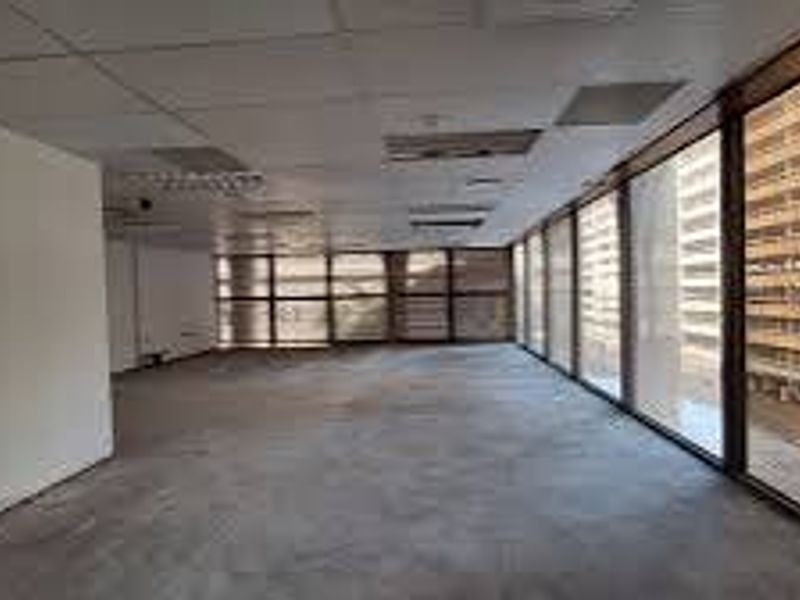 An empty, open-plan commercial office space featuring large windows with city views, a suspended ceiling, and carpeted flooring.