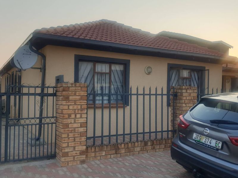 Front elevation of a residential house featuring a tiled roof, brick perimeter wall with metal security fencing, and a paved driveway.