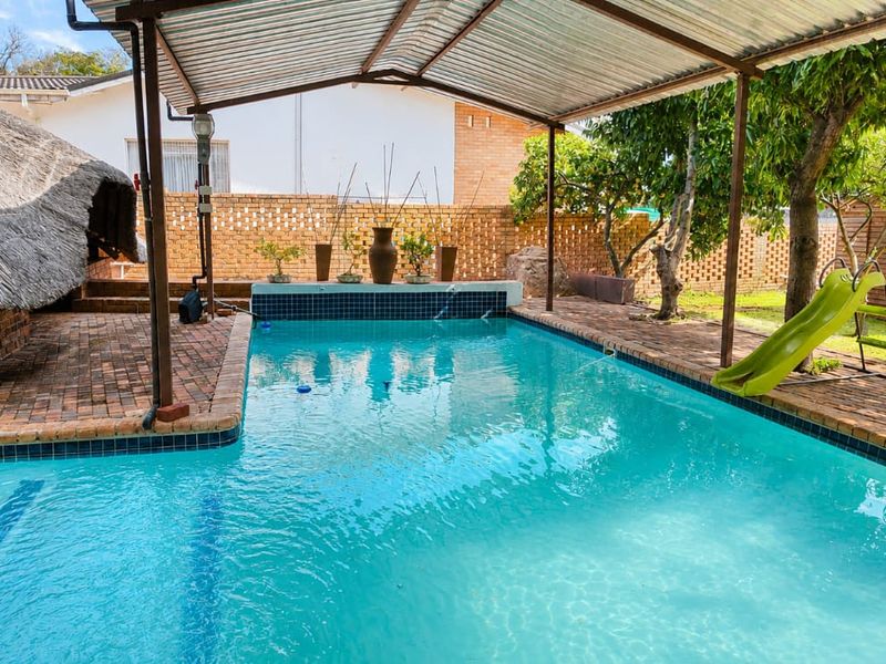 Outdoor pool area featuring a covered patio, brick paving, and a thatched roof structure, set within a private garden with a brick boundary wall.