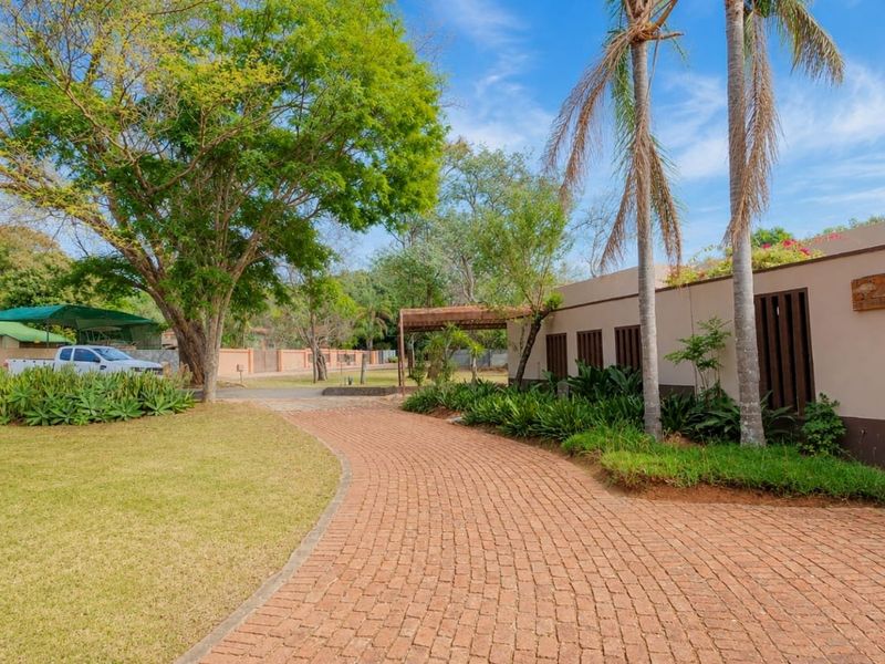 A well-maintained front exterior view featuring a curved brick paved driveway, established trees, and a neat garden leading to the property entrance.