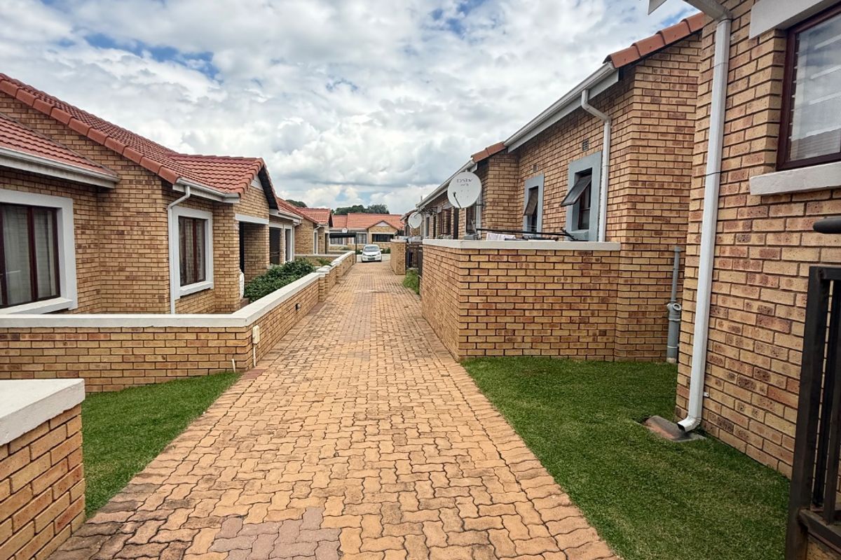 A view of the exterior rear walkway between townhouse units, featuring face brick construction, tiled roofing, and paved pathways with small grass verges.