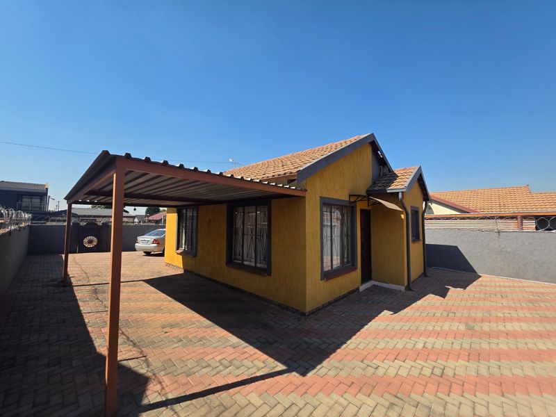 A detached house featuring a yellow textured facade, a covered carport, and a fully paved yard enclosed by perimeter walls with security fencing.