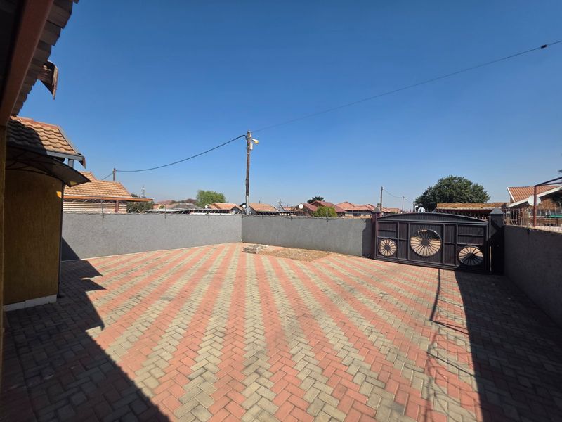 A spacious paved front courtyard featuring a secure metal gate, perimeter walling with security fencing, and clear access to the property entrance.