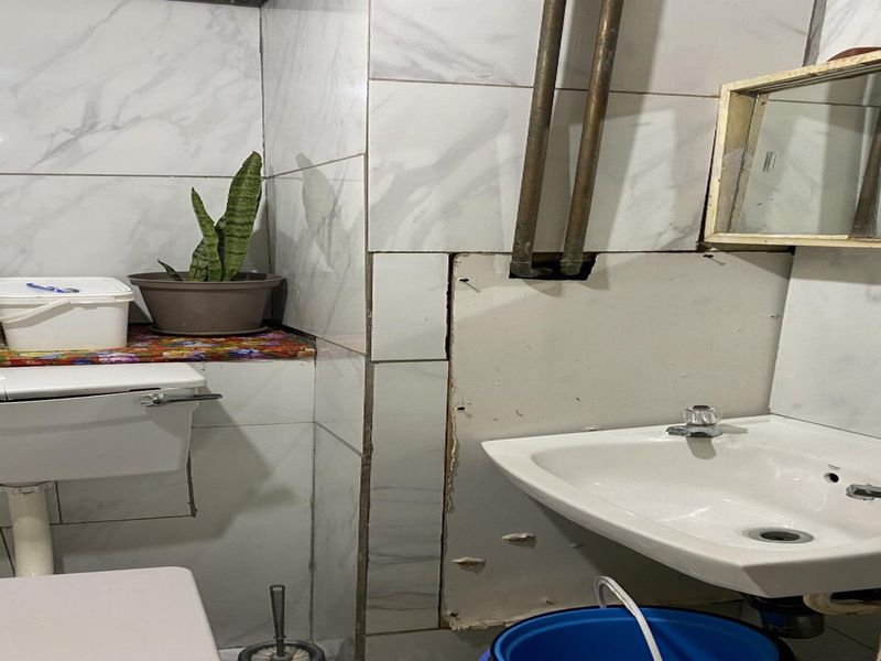 A small bathroom featuring marble-effect wall tiles, a wall-mounted basin, and a toilet, currently showing signs of wear and exposed plumbing infrastructure.