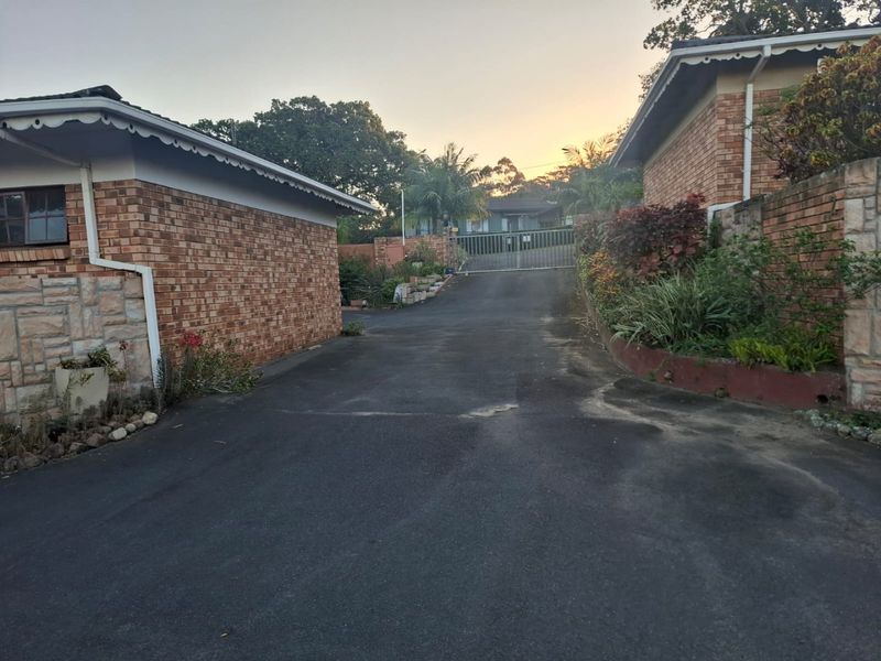 A view of the property driveway leading to a secure metal gate, flanked by brick townhouse structures and landscaped garden beds.