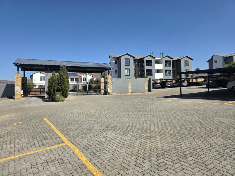 View of the townhouse complex entrance featuring a paved driveway, security gate, and covered parking structures.
