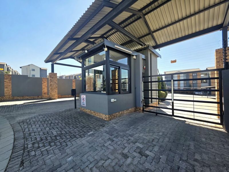 A secure gated entrance to the townhouse complex featuring a guard house, metal sliding gate, and paved driveway with perimeter electric fencing.