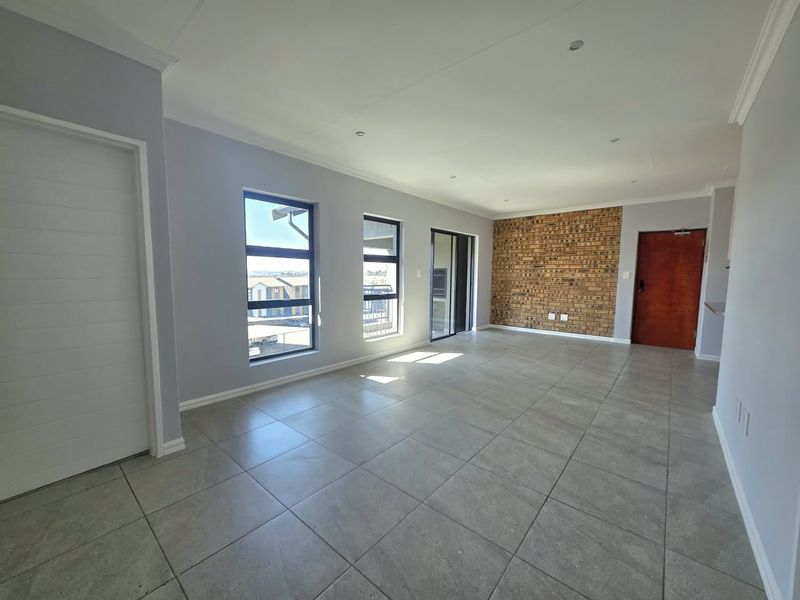 A bright, open-plan living area featuring neutral tiled flooring, a striking exposed brick feature wall, and large windows providing ample natural light.
