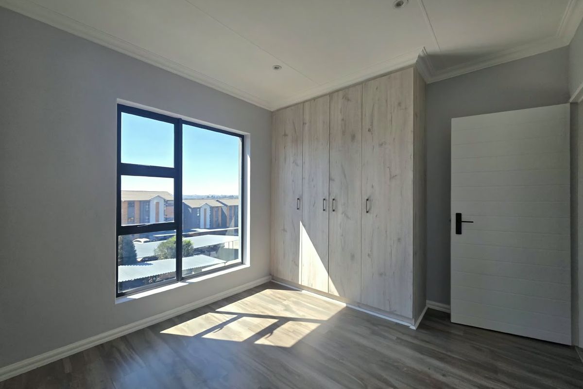 A bright, modern bedroom featuring light wood-finish built-in cupboards, laminate flooring, and large aluminium-framed windows with a view.