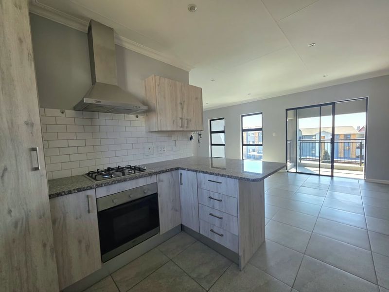 Modern open-plan kitchen featuring light wood cabinetry, granite countertops, and a gas hob, flowing into a bright living area with access to a balcony.