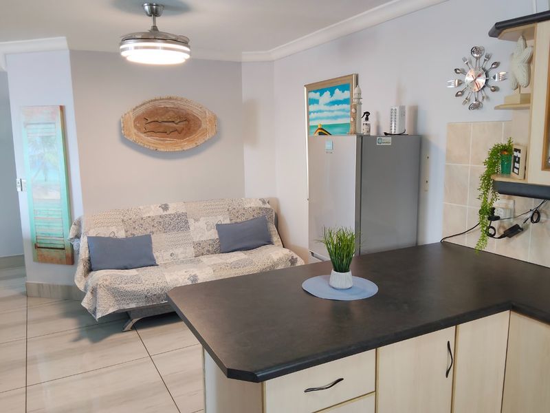 Open plan living area featuring a kitchen with laminate cabinetry and dark countertops, seamlessly flowing into a tiled living space with a ceiling fan.