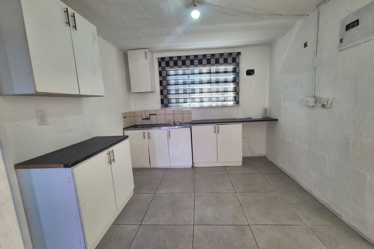 A compact, empty kitchen featuring white built-in cabinetry, dark laminate countertops, tiled backsplash, and grey ceramic floor tiles.