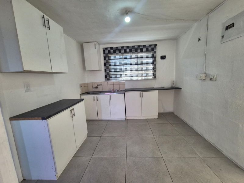 A compact, empty kitchen featuring white built-in cabinetry, dark laminate countertops, tiled backsplash, and grey ceramic floor tiles.