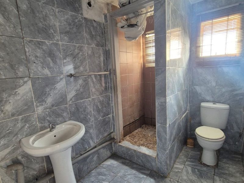 A dated bathroom featuring a pedestal basin, toilet, and a shower enclosure with visible wear and unfinished ceiling details.