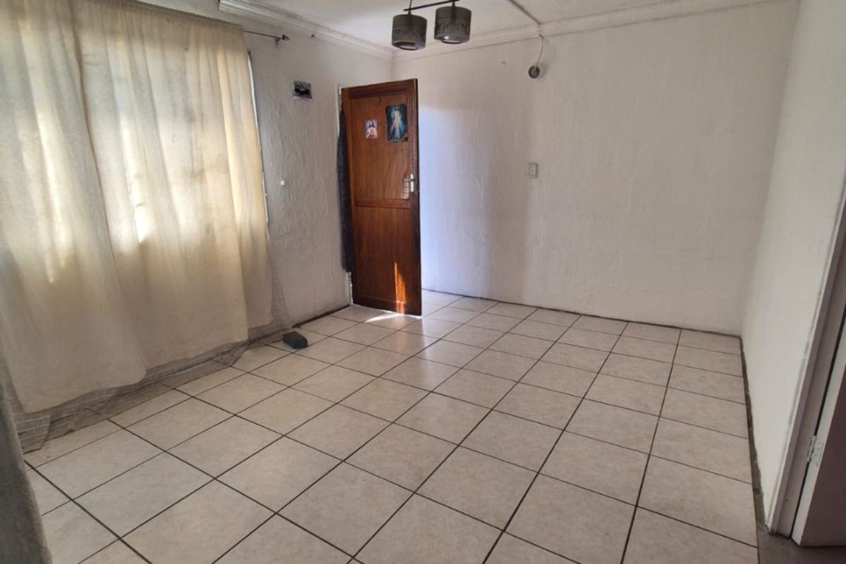 An empty living room featuring ceramic tiled flooring, a ceiling light fixture, and a timber entrance door with natural light from the window.
