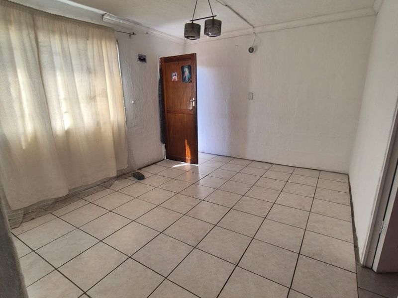 An empty living room featuring ceramic tiled flooring, a ceiling light fixture, and a timber entrance door with natural light from the window.