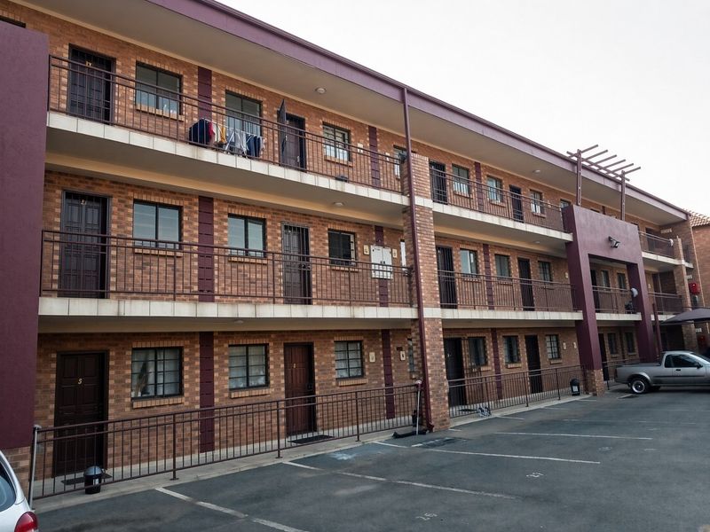 Multi-story apartment building featuring a brick facade, external balcony walkways with metal railings, and an adjacent asphalt parking area.