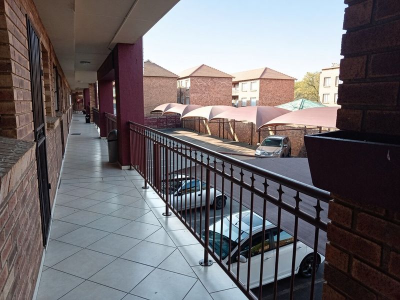 A covered exterior balcony walkway featuring tiled flooring and a metal balustrade, overlooking the apartment complex parking area.