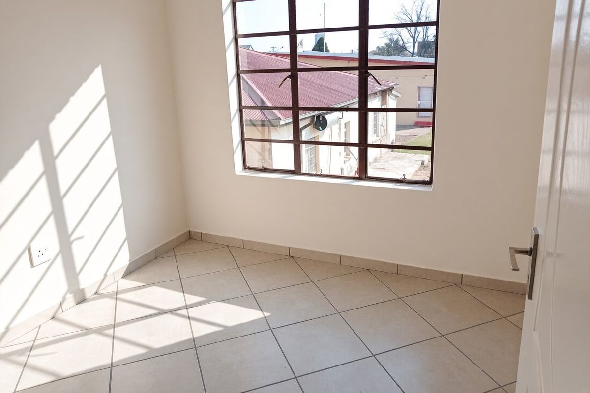 A bright, empty bedroom featuring neutral tiled flooring, clean white walls, and a large steel-framed window providing natural light.
