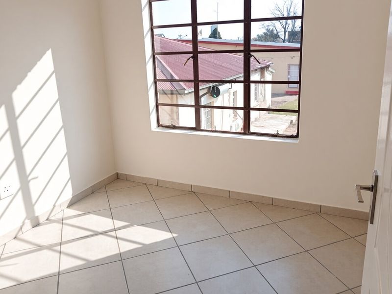 A bright, empty bedroom featuring neutral tiled flooring, clean white walls, and a large steel-framed window providing natural light.