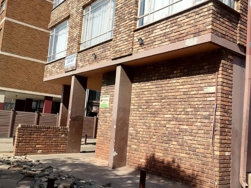 Exterior view of a multi-story face brick apartment building featuring steel frame windows and a paved entrance area with visible maintenance requirements.