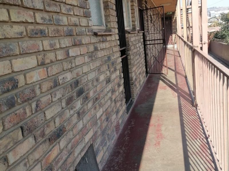 An exterior balcony walkway featuring exposed brick walls, metal security gates, and a metal balustrade overlooking the surrounding area.
