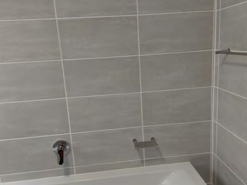 A clean, modern bathroom featuring a white bathtub with chrome fixtures and neutral grey wall tiling.