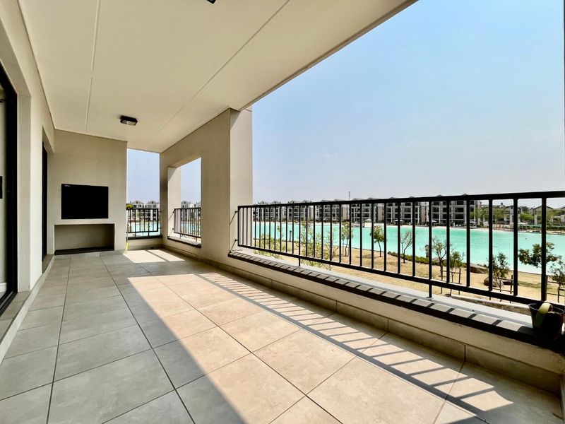 Spacious covered balcony featuring tiled flooring, a built-in braai, and a metal balustrade, offering scenic views of the estate lagoon.