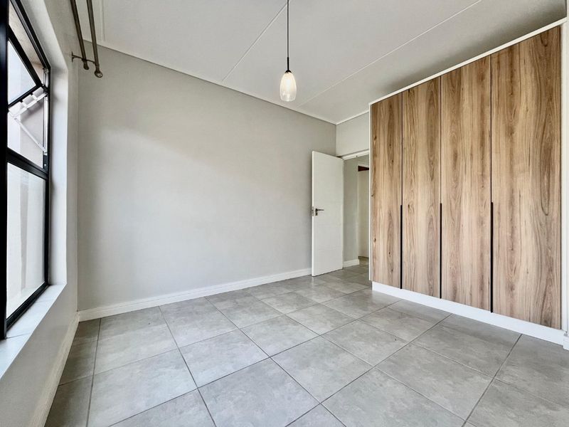 A bright, empty bedroom featuring modern wood-finish built-in cupboards, neutral grey floor tiles, and a clean, minimalist aesthetic.