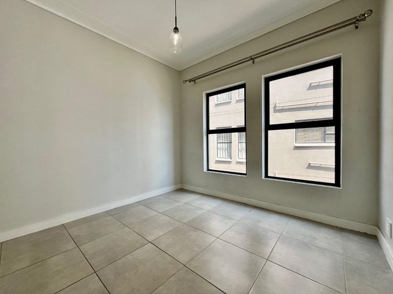 A bright, empty bedroom featuring neutral tiled flooring, modern aluminium window frames, and a simple pendant light fixture.