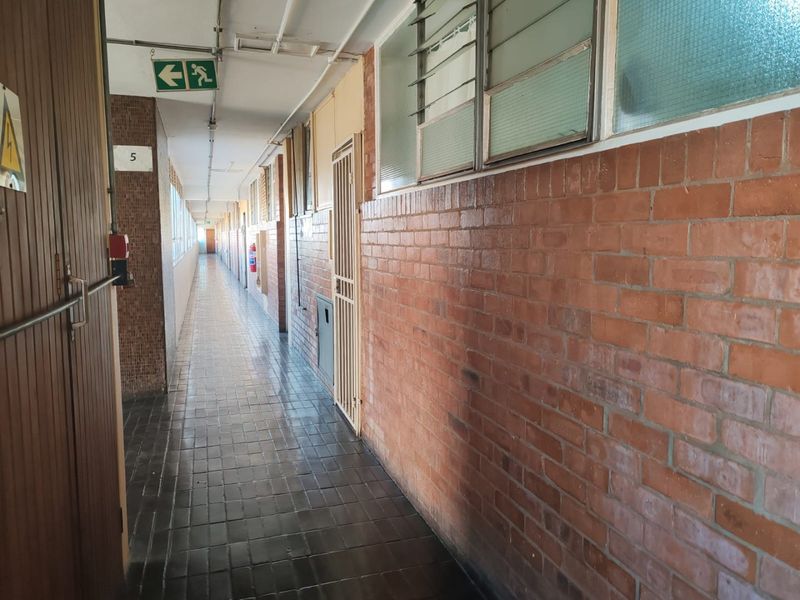 A long, communal apartment hallway featuring exposed brick walls, tiled flooring, and security gates leading to individual units.