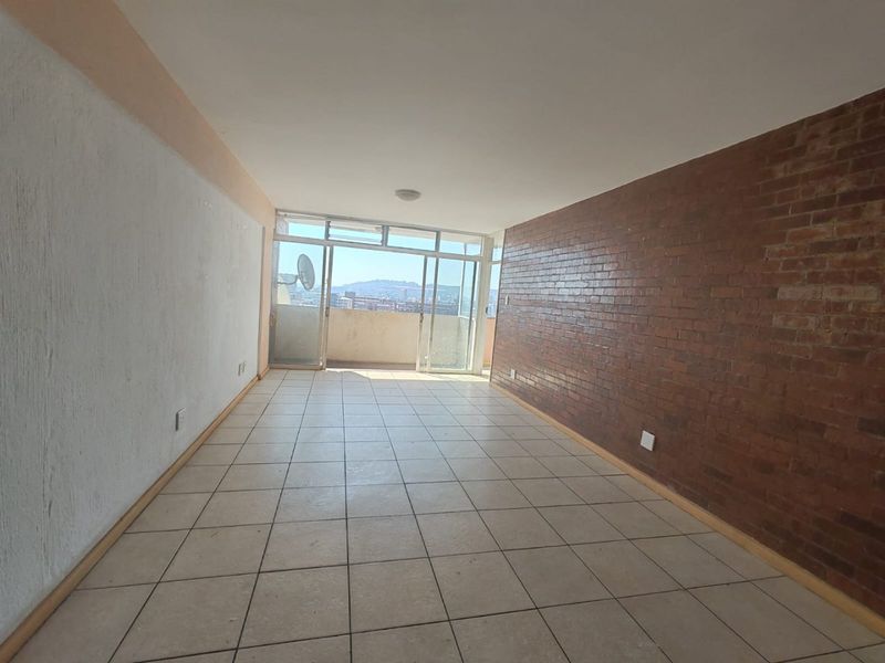 An empty living room featuring an exposed brick feature wall, ceramic tiled flooring, and large sliding glass doors leading to a balcony with city views.