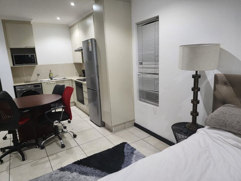 An open-plan studio apartment featuring a compact kitchen with built-in cabinetry, integrated appliances, and tiled flooring throughout the living and sleeping area.