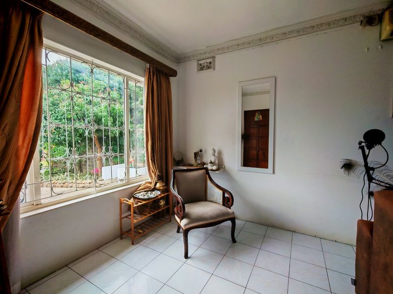 A living area featuring tiled flooring, decorative ceiling cornices, and a large window with security bars overlooking the garden.