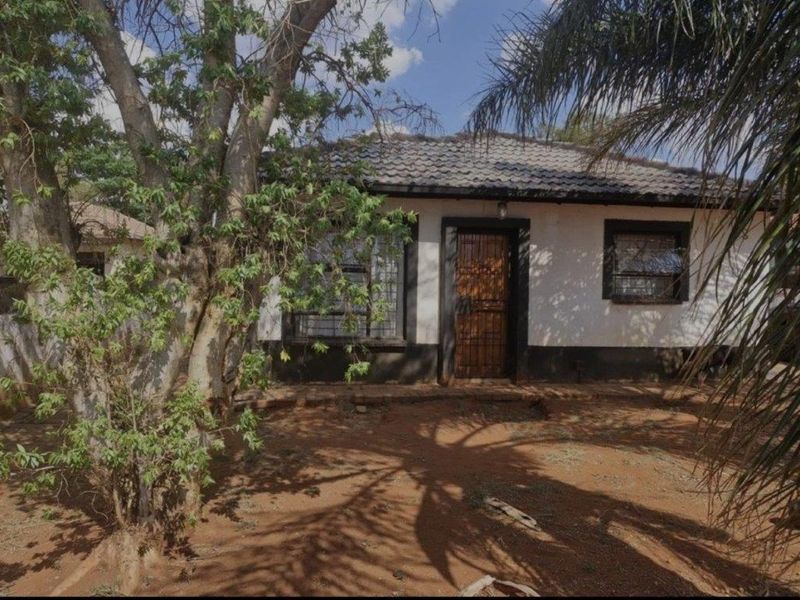 Front elevation of a single-story house featuring a tiled roof, security gate, and burglar bars, set within a natural, unpaved garden area.