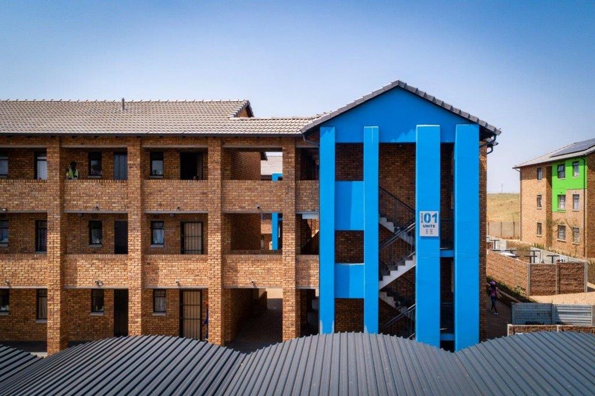 Exterior view of a multi-story apartment block featuring face brick construction, a prominent blue stairwell structure, and corrugated metal roofing.
