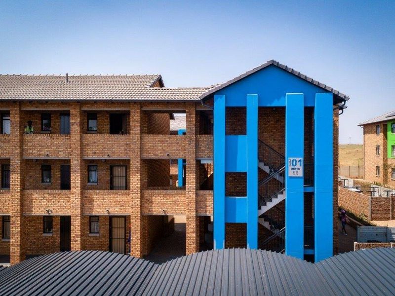 Exterior view of a multi-story apartment block featuring face brick construction, a prominent blue stairwell structure, and corrugated metal roofing.