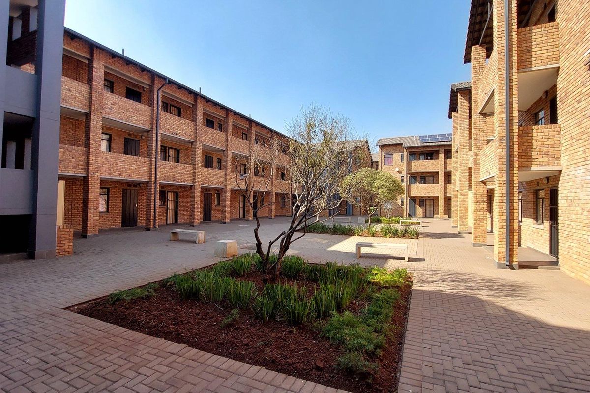 A well-maintained apartment complex courtyard featuring face brick architecture, paved walkways, landscaped garden beds, and communal concrete seating areas.